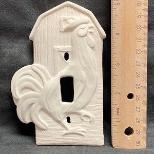 Pode incluir: Uma capa de cerâmica para interruptor de luz em forma de celeiro com um desenho de galo. O galo está em relevo, com cauda e bico detalhados. A capa não está pintada e mede aproximadamente 12.7 cm de altura.