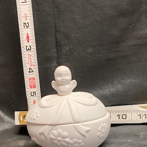 May include: White ceramic egg-shaped trinket box with a lid featuring a small bird figurine and a bow. The box is decorated with floral designs.