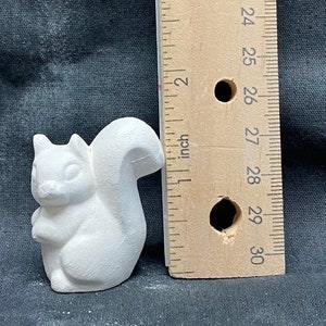 May include: A white ceramic squirrel figurine, approximately 2 inches tall, with a detailed sculpted tail and a cute expression. The figurine is shown next to a ruler for size reference.