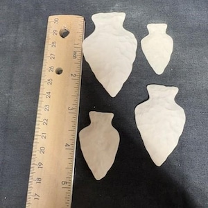 May include: Three off-white, textured arrowhead-shaped objects arranged next to a wooden ruler. The largest arrowhead measures approximately 3 inches in length, while the smaller ones measure approximately 2 inches and 2.5 inches respectively.