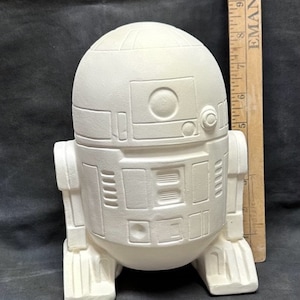 以下が含まれることがあります： 白いセラミック製のR2-D2フィギュア。スターウォーズのキャラクターで、丸い頭と長方形の体です。フィギュアには詳細なパネルと円形の機能があり、背景には定規があります。
