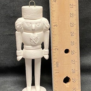 May include: A white ceramic nutcracker figurine, approximately 4 inches tall, with a detailed uniform and a small loop for hanging. The figurine is unpainted and ready for customization.