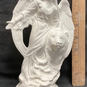 Puede incluir: Figura de cerámica blanca de un ángel con alas, cabello largo y un vestido fluido. El ángel sostiene un cachorro de oso en sus brazos.