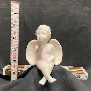May include: A white ceramic cherub figurine, approximately 10 cm tall, blowing a kiss. The cherub has detailed wings and is seated with crossed legs. The figurine is on a dark gray surface.