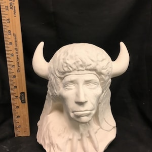 May include: A white ceramic bust of a Native American figure, with a headdress and curved horns, and a detailed facial expression. The bust includes a necklace of three pendant shapes. A ruler is visible on the left side of the image.