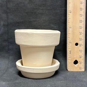 Puede incluir: Una pequeña maceta de terracota sin pintar con un platillo a juego. La maceta mide aproximadamente 7.6 cm de alto, con una superficie ligeramente texturizada. Una regla de madera está al lado de la maceta.