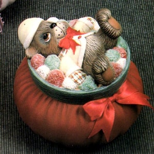 May include: A ceramic Christmas ornament featuring a brown bear wearing a white and red plaid shirt and a white hat, lying in a red sack filled with white and green pom-poms. The sack has a green rim and a red bow.