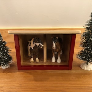 May include: A miniature red and wood-toned horse stable with two toy horses inside. Two small, snow-covered evergreen trees flank the stable. The horses are a brown and white paint and a brown and white Clydesdale.