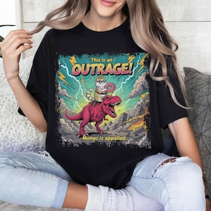 May include: Black t-shirt featuring a colorful graphic of a cat riding a pink dinosaur with the text "This is an OUTRAGE!" and "Mongo is appalled." The design includes lightning bolts, clouds, and a cityscape.