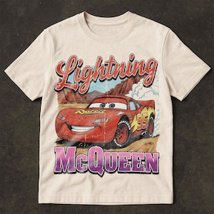 May include: Cream-colored t-shirt featuring a distressed graphic of Lightning McQueen, a red race car with the number 95. The text "Lightning McQueen" and "95" are prominently displayed in red and yellow, with the name "McQueen" in purple.