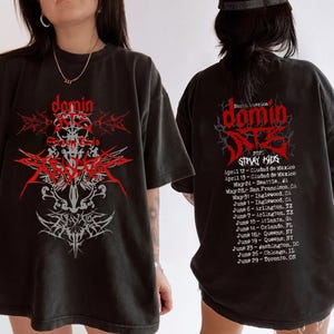 May include: Black t-shirt featuring a red and gray graphic design with the word "domin" and the text "STRAY KIDS 2025" on the front. The back of the shirt displays a tour schedule. The shirt has short sleeves and a relaxed fit.