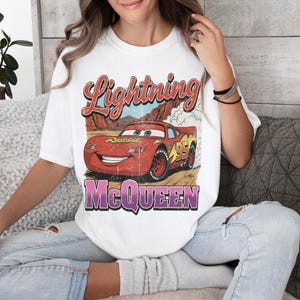 May include: White t-shirt featuring a graphic of Lightning McQueen from the Cars movie. The design includes the text "Lightning McQueen" in red and purple, and the number "95" on the car. The shirt has a vintage, distressed look.