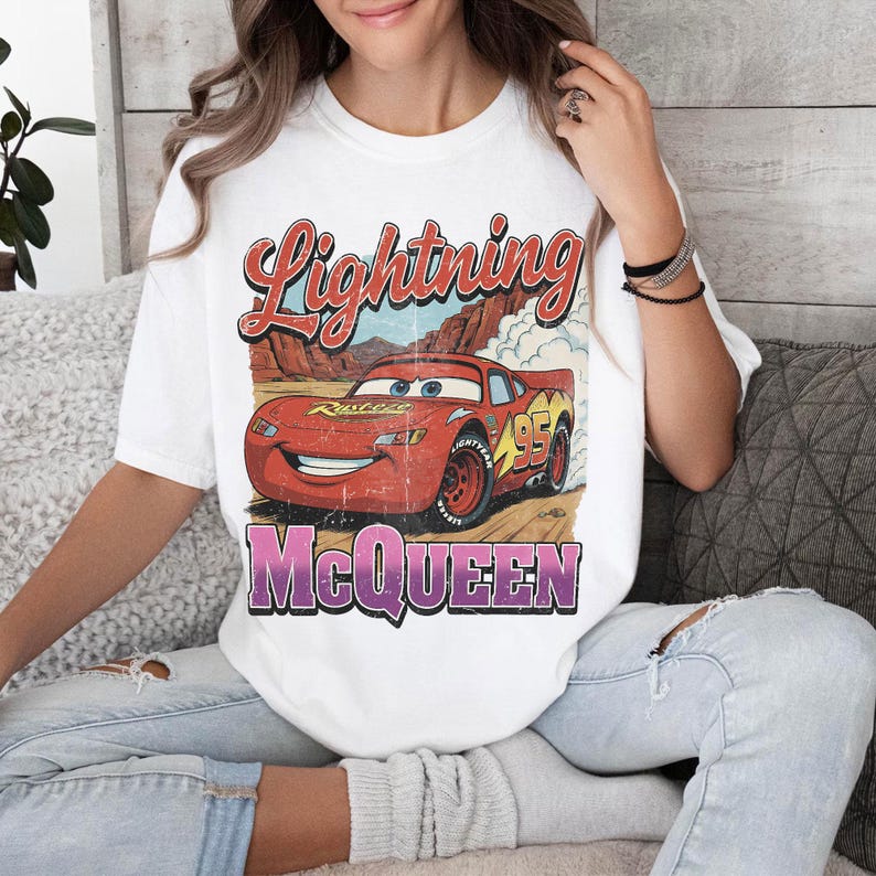 May include: White t-shirt featuring a graphic of Lightning McQueen from the Cars movie. The design includes the text "Lightning McQueen" in red and purple, and the number "95" on the car. The shirt has a vintage, distressed look.