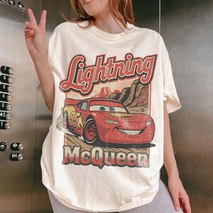 May include: Cream-colored t-shirt featuring a graphic of Lightning McQueen from the movie Cars. The design includes the text "Lightning McQueen" in red and the number "95" on the car. The shirt has a vintage, distressed look.