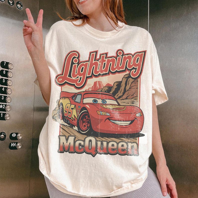 May include: Cream-colored t-shirt featuring a graphic of Lightning McQueen from the movie Cars. The design includes the text "Lightning McQueen" in red and the number "95" on the car. The shirt has a vintage, distressed look.