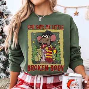 May include: Olive green sweatshirt featuring a cartoon rat character wearing a hat and scarf, framed by Christmas trees. The text "GOD SAVE MY LITTLE BROKEN BODY" is printed above and below the image in red and gold.