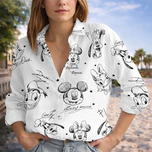 Puede incluir: Camisa blanca abotonada con bocetos en blanco y negro de personajes de Disney como Mickey Mouse, Minnie Mouse y Donald Duck. La camisa tiene mangas largas y cuello. Los nombres de los personajes están escritos en cursiva.