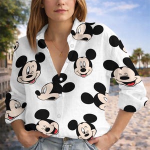 Puede incluir: Camisa blanca con botones con un estampado repetido de caras de Mickey Mouse en blanco y negro. La camisa de manga larga tiene cuello y está hecha de una tela ligera. La camisa está desabrochada en la parte superior.