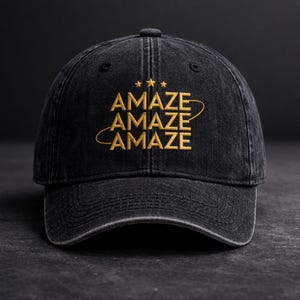 Könnte beinhalten: Schwarze Baseballkappe aus Denim mit dem Wort "AMAZE" in Gold gestickt. Das Wort wird dreimal wiederholt, mit drei kleinen Sternen über dem oberen Wort. Die Kappe hat einen gebogenen Schirm und einen Stoffriemenverschluss.