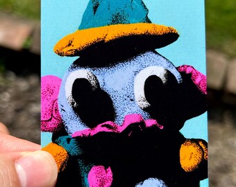 May include: A rectangular print featuring a stylized character with a light blue head, large black eyes, and a teal hat with an orange brim. The character has a dark body accented with bright pink and orange polka dots against a light blue background.