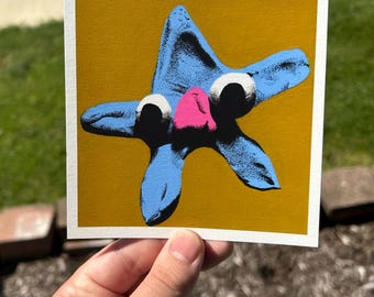 May include: A blue starfish illustration with a pink nose and white eyes on a mustard yellow background. The artwork is on a white square card. The image is held by a hand.