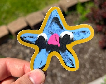May include: A die-cut sticker in the shape of a blue starfish with a cartoon face. The starfish has a pink mouth, white eyes, and a yellow border. The sticker is held in a hand against a blurred outdoor background.