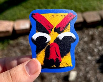 May include: A rectangular sticker with a blue border featuring a stylized graphic design. The center is yellow with two red diagonal shapes at the top, two large white circular eyes with black pupils, and a red nose shape in the lower center.