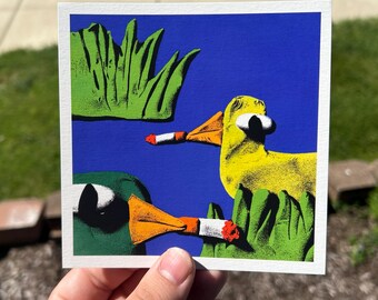 May include: A square art print featuring two cartoon ducks with cigarettes in their beaks. One duck is yellow, the other is green. The background is blue with green grass.