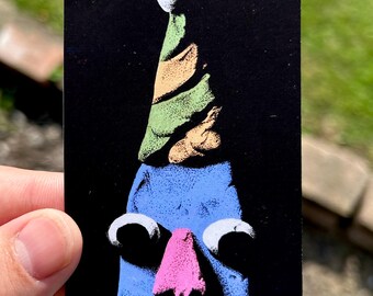 May include: A rectangular sticker featuring a stylized blue character against a black background. The character wears a green and orange striped party hat with a white pom-pom, has two large white circular eyes, and a small pink triangular nose.