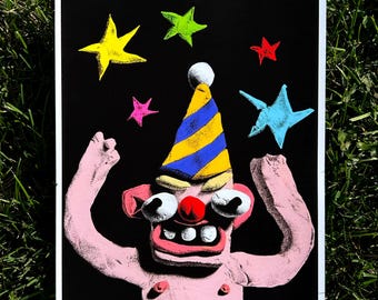 May include: A rectangular art print on a black background featuring a pink, clay-like figure with a red nose, large white eyes, and a wide mouth with teeth. The figure wears a yellow and blue striped party hat and is surrounded by five colorful stars.
