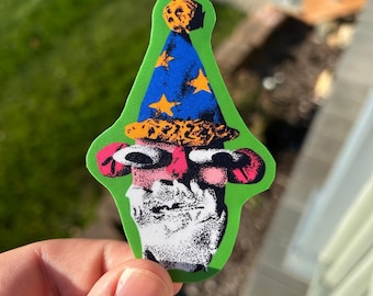May include: A die-cut sticker featuring a stylized character with a white beard, pink cheeks, and wide eyes. The character wears a blue cone-shaped hat decorated with yellow stars and a textured orange brim, all set against a bright green background.