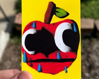 May include: A rectangular sticker with a bright yellow background featuring a stylized red apple illustration. The apple has large white eyes, a black mouth, and several blue teardrops falling from its eyes and around its red body.