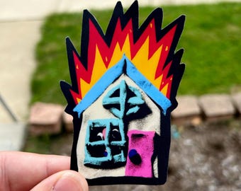 May include: A die-cut sticker featuring a stylized house engulfed in bright yellow and red flames. The house has a light-colored facade, a blue roof, a blue window frame with a cross design, and a vibrant pink door with a dark blue circular knob.
