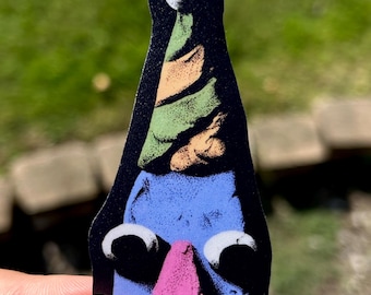 May include: A die-cut sticker featuring a blue character with two large white eyes and a pink nose. The character wears a tall, pointed party hat with alternating green and tan diagonal stripes and a white pom-pom at the very top, all on a black background.