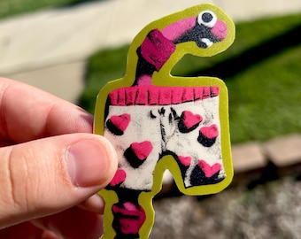 May include: A die-cut sticker featuring a cartoon worm with a pink and black body wearing white boxer shorts decorated with a pattern of small pink hearts. The sticker has a thick, bright lime green border outlining the entire character shape.