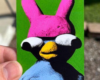 May include: A rectangular sticker featuring a stylized character with a bright pink hat with two tall ears, large white eyes, a yellow beak, and a light blue body, all set against a solid, vibrant green background with a grainy, textured print effect.