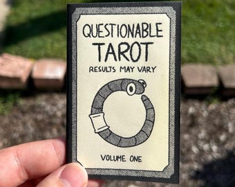 May include: A rectangular card with a cream-colored center and a black border. The text reads QUESTIONABLE TAROT, RESULTS MAY VARY, and VOLUME ONE. A black and white illustration of a worm curled into a circle is centered on the card.