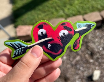 May include: A die-cut sticker featuring a red heart with a cartoon face and wide eyes, pierced by a black and white arrow. The heart has a thick, bright green border and a small red droplet extending from the bottom right side of the heart shape.
