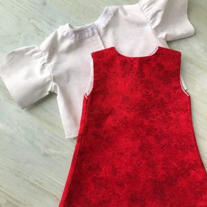 May include: A white top with short sleeves and a lace collar, alongside a red dress with a mottled pattern. The garments are likely for a doll or small child. The items are made of fabric.