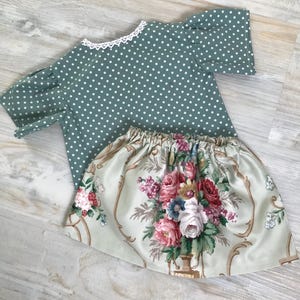 May include: A two-piece outfit. The top is sage green with white polka dots and a white lace trim. The skirt has a floral pattern with pink, red, and blue flowers on a light green background.