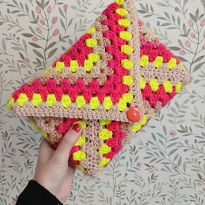 Peut inclure: Pochette au crochet faite à la main, motif granny square. La pochette présente des carrés rose vif, jaune fluo et beige, avec une bordure beige et une fermeture à bouton orange vif. La pochette est tenue dans une main.