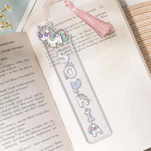 Op de afbeelding: Transparante acryl boekenlegger met de naam "SOPHIA" in kleurrijke, grillige letters en eenhoorn illustraties. De boekenlegger heeft een roze kwast en ligt op een open boek. Een uniek cadeau voor boekenliefhebbers.