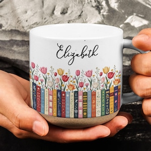 May include: A white ceramic mug featuring a row of colorful book spines at the base, topped with a vibrant illustration of various tulips and wildflowers. The name Elizabeth is printed in black cursive script above the floral design.