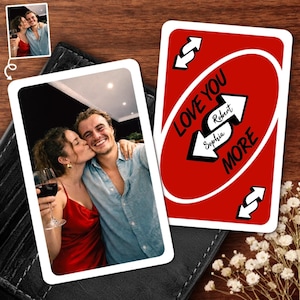 Puede incluir: Una tarjeta fotográfica personalizada y una tarjeta roja con las palabras "LOVE YOU MORE" y los nombres "Robert" y "Sophia" impresos. La tarjeta fotográfica muestra a una pareja abrazándose y besándose. Las tarjetas están sobre una superficie de madera.