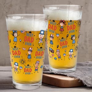 May include: Two clear pint glasses filled with beer and foam. The glasses are decorated with cartoon illustrations of children and the text "BEST DAD EVER" in red and yellow. The glasses are on a wooden surface.