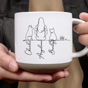 May include: White ceramic mug with a brown base, featuring a line art illustration of a person, a dog, and a cat. The names "Charlie", "Angelina", and "Barley" are written below the figures.