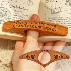 May include: A wooden thumb page holder with the text "I love you to the moon and back HENRY" engraved on it. The page holder is in use, holding open a book. A second page holder with a moon phase design is also visible.
