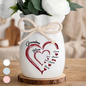 May include: White ceramic vase with a heart design and the words "Grandma" and "Mom" in black script. The vase has a rope bow and is decorated with red hearts and names. The vase is sitting on a wooden coaster and contains white flowers.