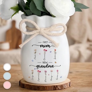 May include: White ceramic vase with a cream-colored rope bow, holding white flowers. The vase is decorated with the text "First Mom Now Grandma" and floral illustrations with names. It sits on a wooden coaster.