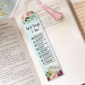 Könnte beinhalten: Ein floraler Lesezeichen mit einer rosa Quaste, auf dem der Text "Gott sagt, ich bin" und eine Liste von Affirmationen steht. Das Design umfasst Aquarellblumen und einen hellblauen Hintergrund. Das Lesezeichen liegt auf einem offenen Buch.